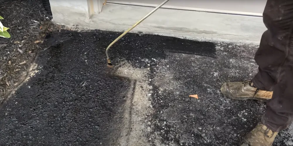 Vue rapprochée de l'application d'un scellant sur une vieille entrée en asphalte, avec le spray qui rend la surface humide et sombre